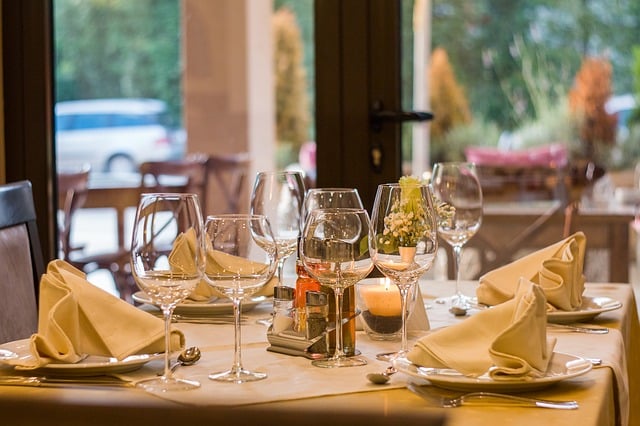 A restaurant table set for four with folded beige serviettes, wine glasses, a small candle, cutlery, and a flower arrangement—an inviting spot ideal for a special birthday meal, with outdoor seating visible through the window in the background.