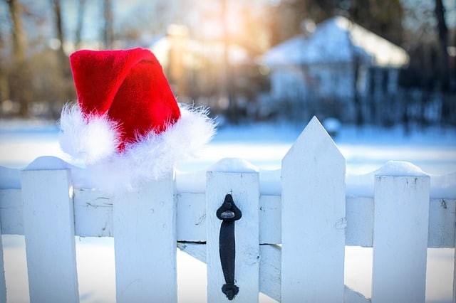 A red Santa hat with white trim rests on a white wooden fence. Snow covers the ground, evoking a bright Lapland day, with houses and trees blurred in the background—a magical winter scene perfect for children.