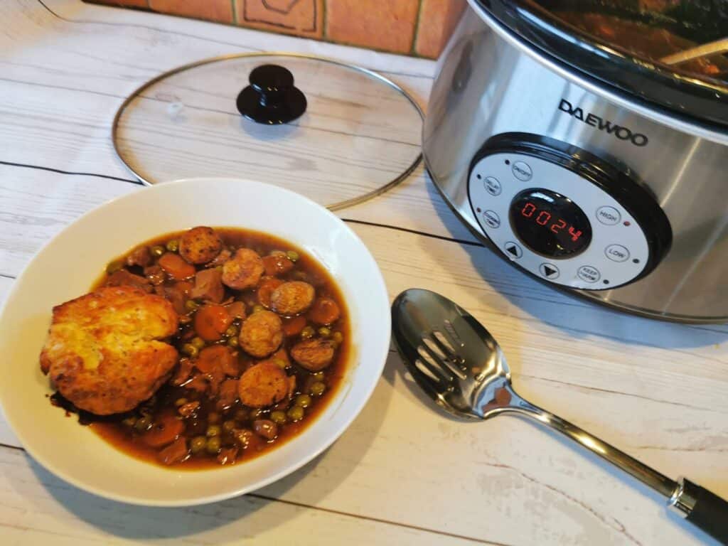 A bowl of stew with vegetables, sausage slices, and a scone sits beside a spoon and a Daewoo slow cooker on a light wooden surface—one of those family favourite meal ideas perfect for sharing. The cooker's lid is off, revealing food inside; its timer reads 00:24.
