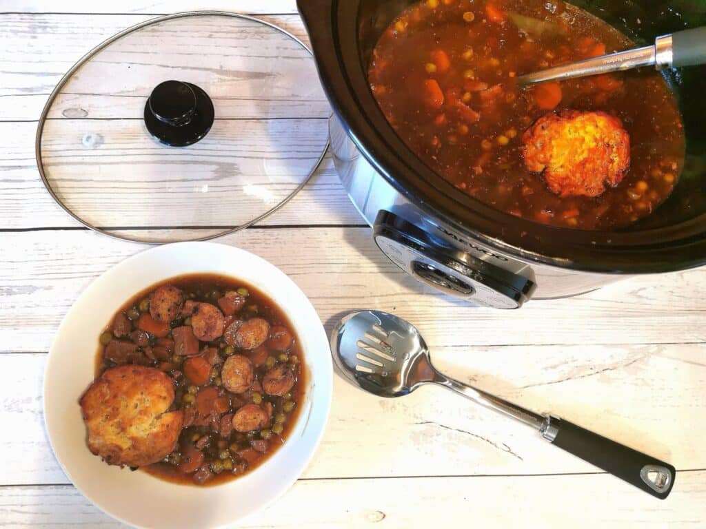 A bowl of stew with vegetables, sausage slices, and a scone on top sits next to a slow cooker filled with more stew and a ladle, on a white wooden surface—a perfect addition to your family favourite meal ideas. A slotted spoon is nearby.