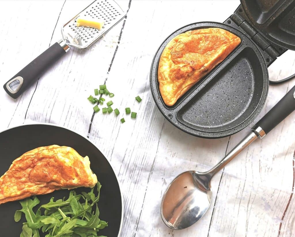 A folded omelette in a sandwich maker sits open next to a spoon and cheese grater on a white wooden surface. A plate with rocket and another omelette is nearby, making this scene perfect for family favourite meal ideas. Chopped chives are scattered around.