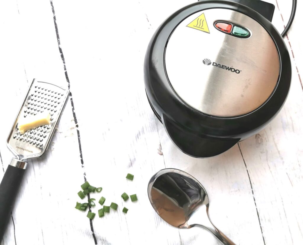 A Daewoo waffle maker sits on a white wooden surface next to a metal spoon, a grater with cheese, and scattered chopped spring onions—perfect for trying out new family favourite meal ideas.