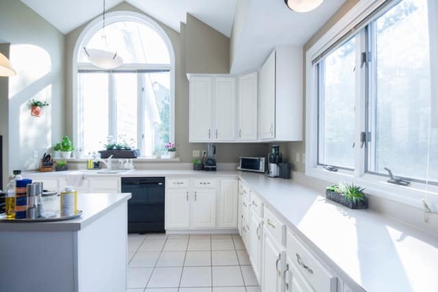 Bright, modern kitchen with white cupboards and large windows letting in natural light—perfect for those seeking tips for lighting or bright ideas on lighting a small room. Black appliances, houseplants, and a kitchen island complete the inviting space.