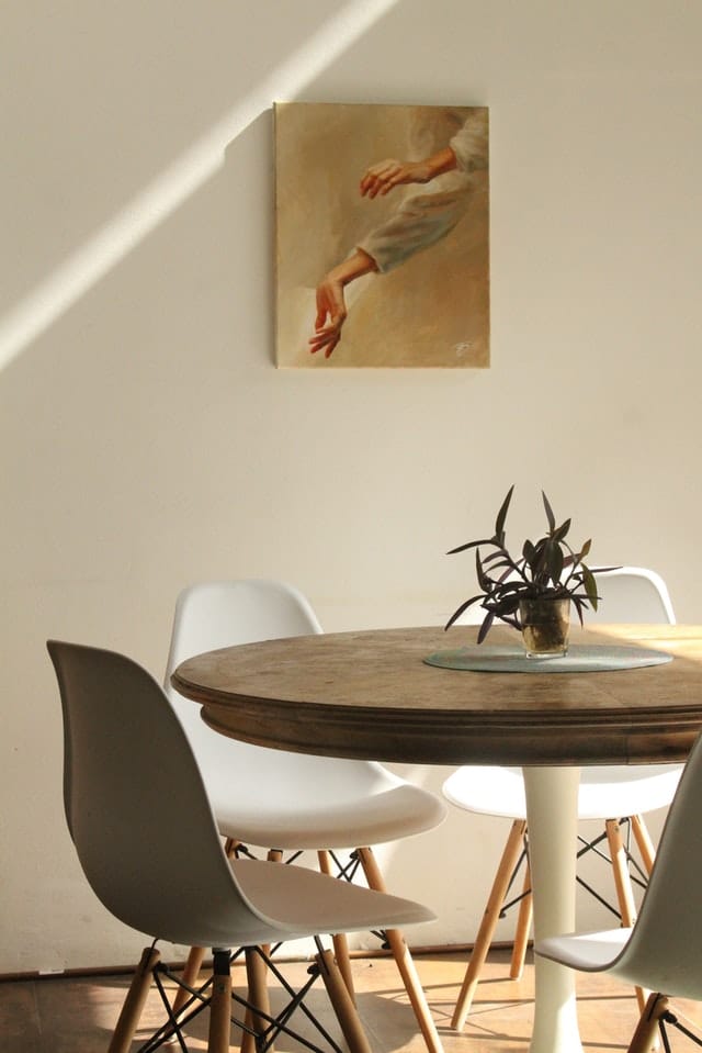 A round wooden table with four white chairs sits in natural sunlight, offering bright ideas for lighting a small room. A small potted plant graces the table, and a painting of hands and arms hangs on the plain white wall behind.