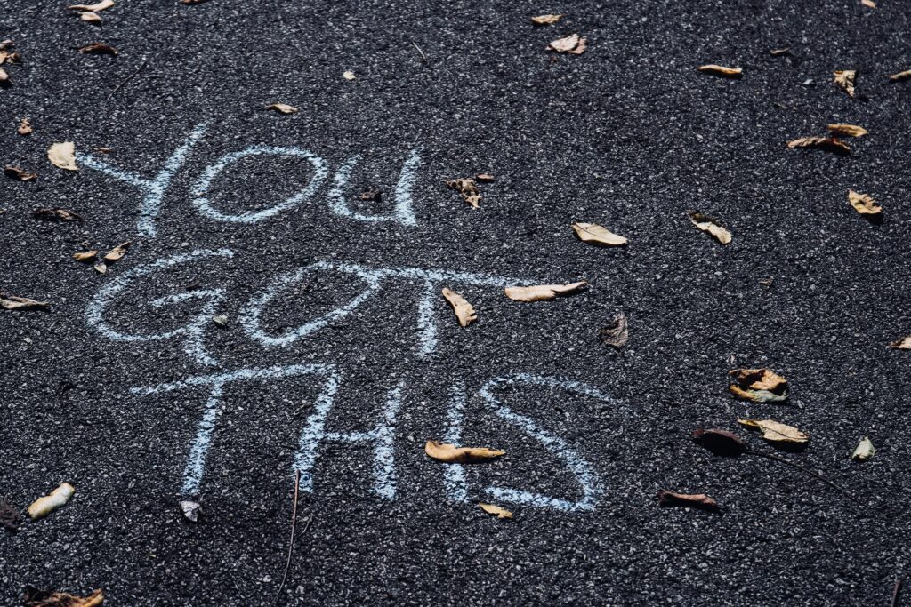 White chalk message "YOU GOT THIS" is written on a dark tarmac surface, surrounded by scattered dried leaves—a simple reminder and one of those self-esteem boosting tips you might find on a walk.