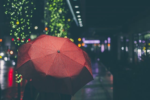 A person holds a red umbrella on a rainy city street at night, the blurred city lights creating a festive atmosphere—perfect reminder to learn some umbrella tips so you stop forgetting your umbrella on nights like this.