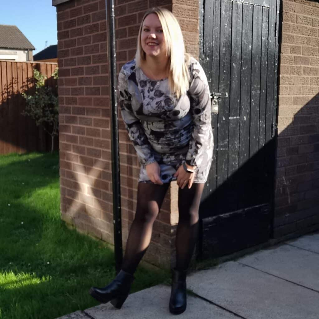 A woman with blonde hair, wearing a patterned dress, black tights, and black boots, stands smiling near a brick shed in a sunny garden—ready to win a £20 Amazon Voucher.