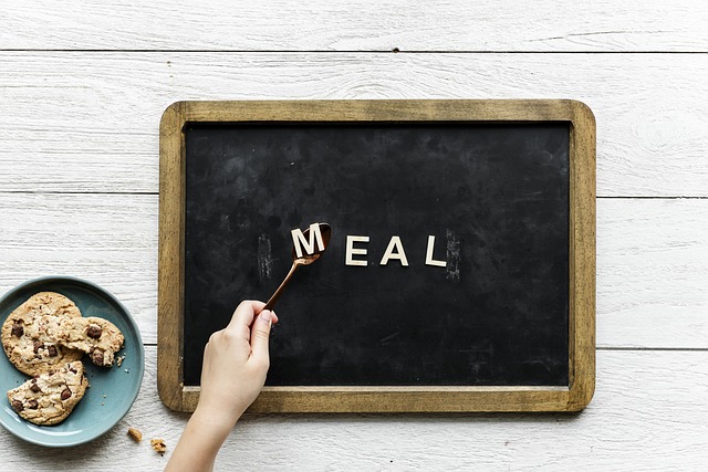 A child’s hand uses a fork to move the letter “M” on a chalkboard spelling “MEAL,” offering quick tips to make your meals more exciting. Beside the board sits a plate of chocolate chip biscuits on a white wooden surface.