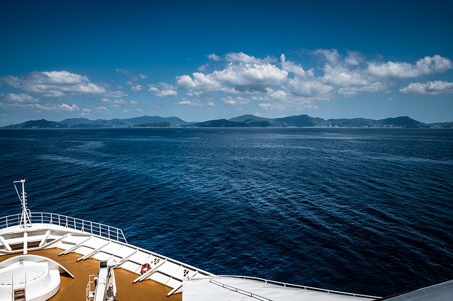 The image shows the bow of a ship sailing on calm, deep blue water under a partly cloudy sky, with a distant green mountain range on the horizon—a scene reminiscent of popular cruise destinations.