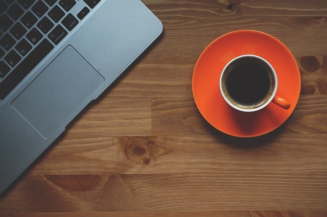 A silver laptop sits on a wooden table next to a cup of black coffee in an orange saucer—the perfect setup for brainstorming clever business ideas, viewed from above.