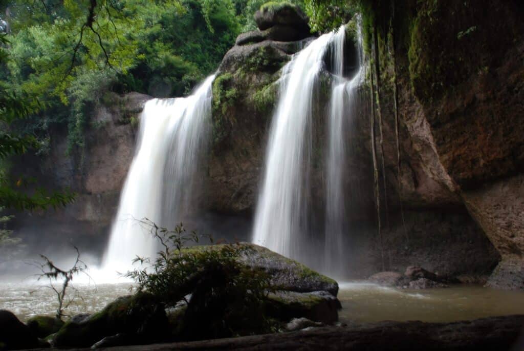 A wide waterfall cascades over a rocky cliff surrounded by lush green foliage, with mist rising from the water below and moss-covered rocks in the foreground—a breathtaking scene straight from any film travel bucket list.