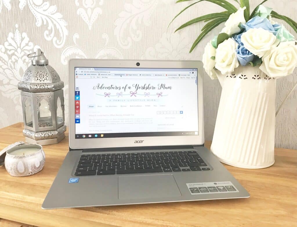 A laptop sits on a wooden desk, displaying a blog about starting an online business. Next to it are a white jug with blue and white flowers, a decorative lantern, and a lit candle against a grey patterned wall.