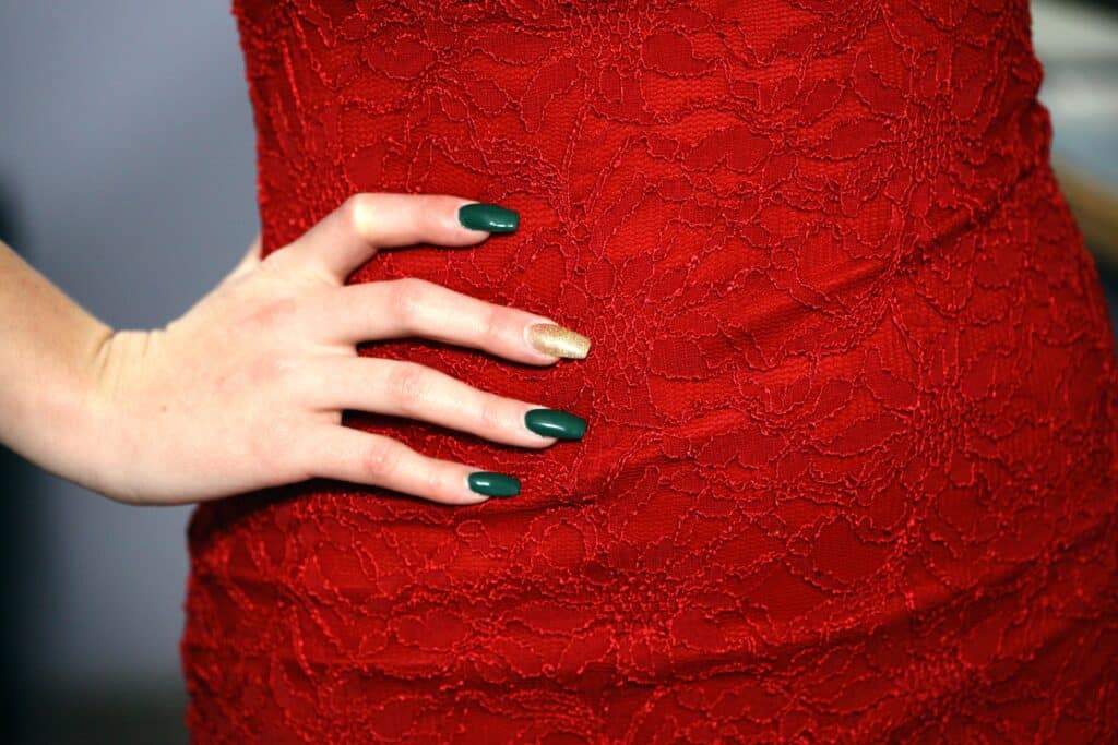 A hand with long nails painted green, except for one gold accent nail, rests on the hip of a person wearing a fitted, red lace bodycon dress.