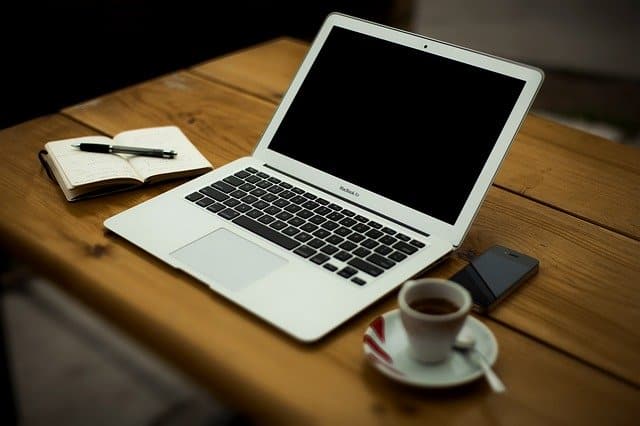 A silver laptop on a wooden table next to an open notebook with a pen, a mobile phone, and a cup of coffee on a saucer—perfect for jotting down self-employment tips or important things to consider for the self-employed.