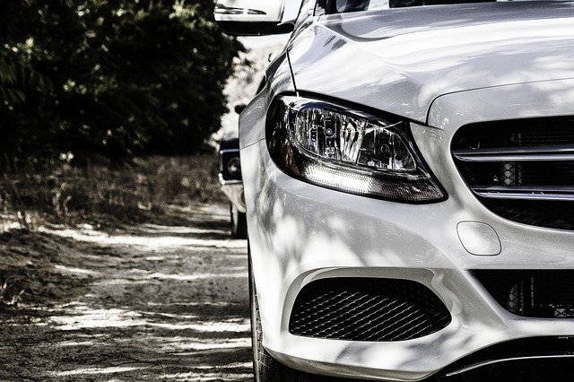Close-up of the front right side of a white car with private number plates, parked on a dirt road, another car behind it and trees in the background.
