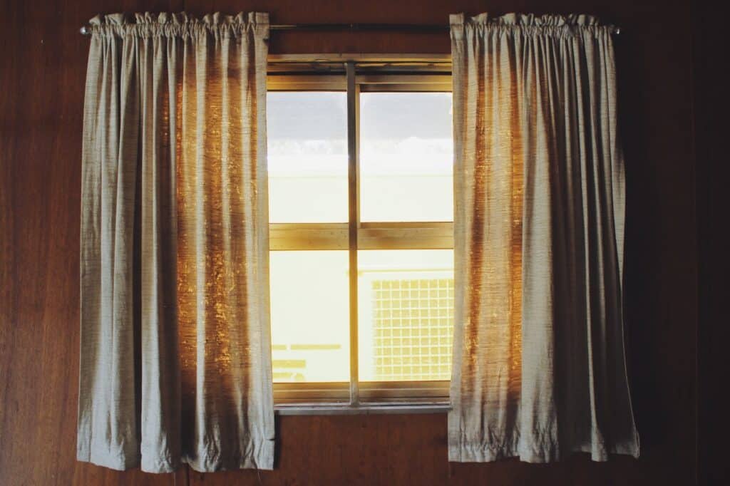 A wooden-framed window with sheer beige curtains partially drawn open allows warm sunlight to filter through—one of the simple ways to increase natural light in your home. The wooden-panelled walls create a cosy, vintage atmosphere.