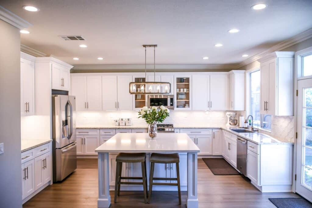 Bright, modern kitchen with white cupboards, stainless steel appliances, and a centre island with two chairs—simple ways to make your home feel open. Large windows and recessed lighting increase natural light throughout the space.
