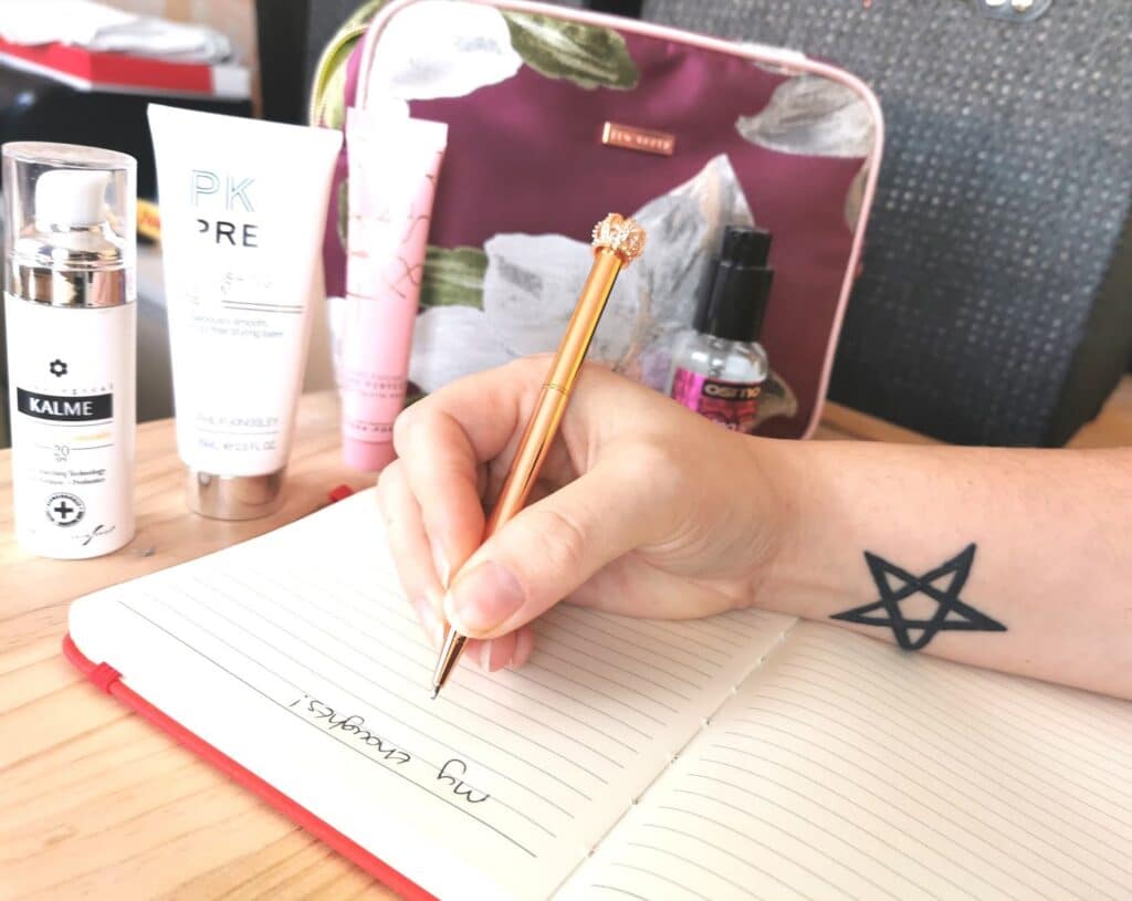 A person with a pentagram tattoo on their wrist writes “my essentials” in a notebook. Skincare products and a floral make-up bag hint at product testing on the wooden table in the background.