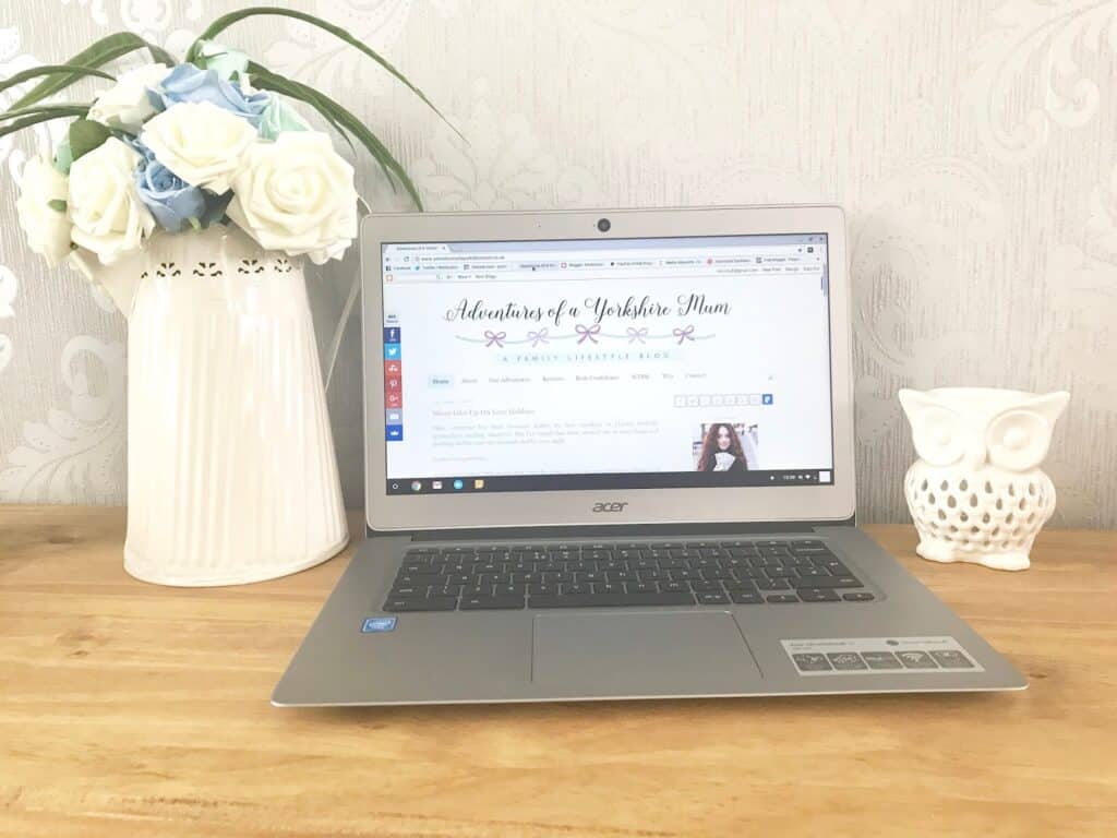A silver laptop displaying a blogging site sits on a wooden desk next to a white vase with blue and white flowers and a white ceramic owl ornament, against a light-patterned wallpaper background.