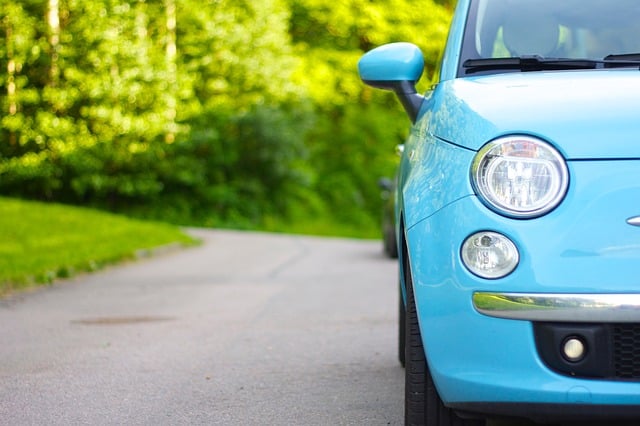 Close-up of the front right side of a light blue Fiat 500 parked on a road, with lush green trees and grass in the background. The bright, sunny scene perfectly captures the charm that fuelled the Fiat 500 rise.