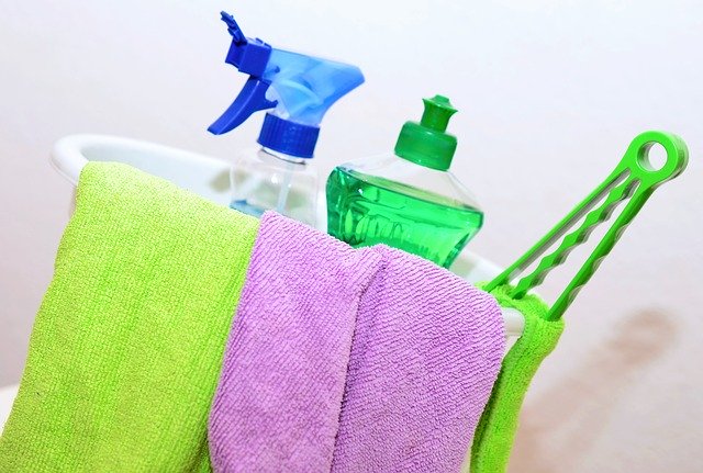 A white cleaning caddy holds a green washing-up liquid bottle, a blue spray bottle, a green cleaning brush, and green and purple towels draped over the side—essential tools to clean your home like a professional cleaning expert.