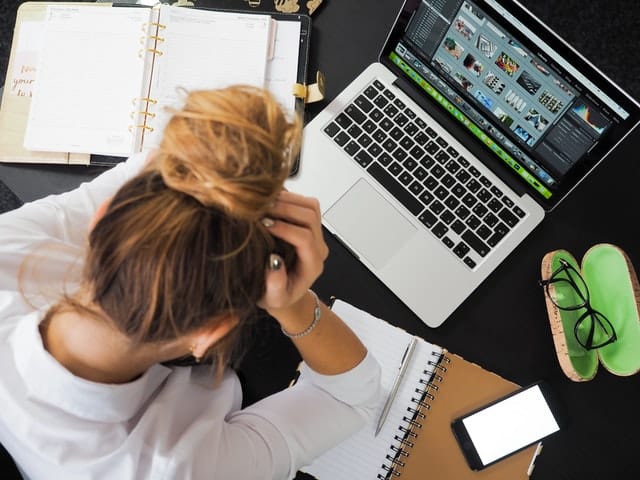 A person sits at a desk with their head in their hands, surrounded by a laptop, notebooks, a mobile phone, spectacles, and papers—capturing workplace stress in a challenging work environment.