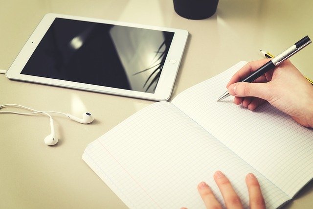 A person writes in a notebook with a pen at a desk, possibly seeking child homework help. An open tablet, white earphones, and a cup are also on the desk, suggesting a focused environment for studying or helping children with homework.