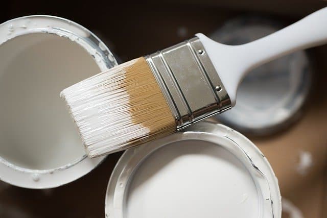 A paintbrush with white paint on its bristles rests across the open top of a white paint tin, while another similarly open tin sits nearby—an inviting start for anyone wondering how to paint your kitchen cupboards.