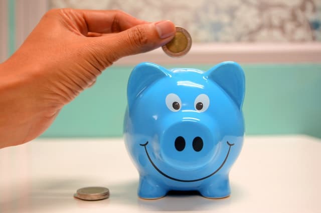 A hand is placing a coin into a smiling blue piggy bank on a white surface, showing simple ways to save for the New Year, with a few other coins nearby.