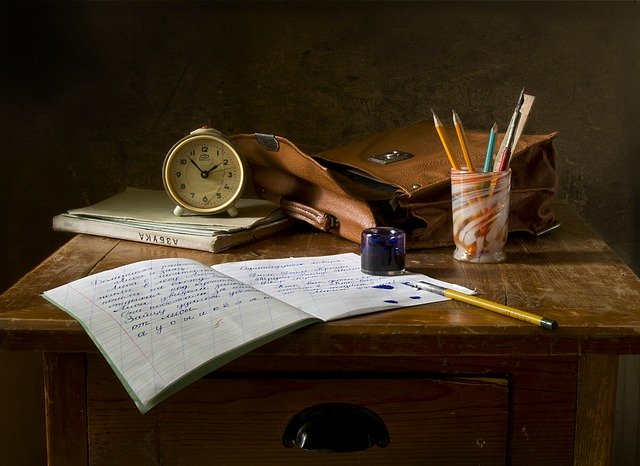 An open notebook with handwritten text sits on a wooden desk beside supplies, evoking tips for going back to school—complete with a pencil, ink jar, alarm clock, closed book, cup of pens and pencils, and a brown leather satchel.