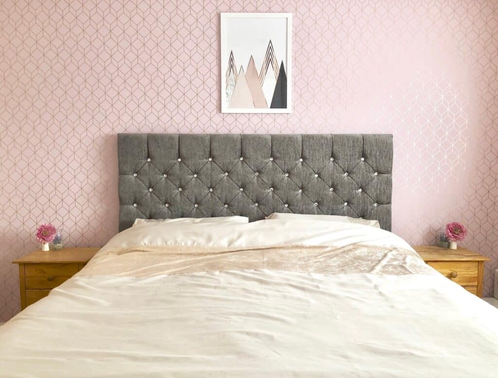 A neatly made bed with a tufted grey headboard anchors this modern master bedroom, flanked by wooden bedside tables with pink flower arrangements. Pink patterned wallpaper and a geometric mountain art print add fresh modern colour ideas.