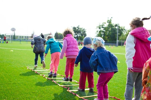 A group of young children in colourful jackets walk in a line, stepping over a rope ladder during PE on green grass at an outdoor sports field, promoting physical activity and mental health on a sunny day.