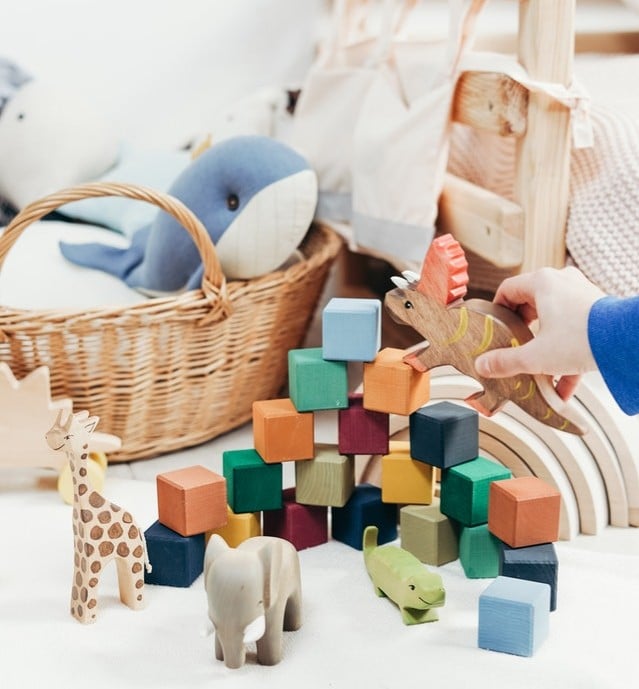 A child’s hand holds a wooden dinosaur toy above a colourful stack of blocks. Other wooden toys rest near a wicker basket with a soft toy whale, showcasing playroom organisation that makes tidying up easy and keeps the space tidy.