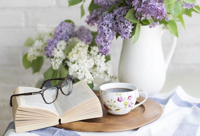 An open book—one of the classic novels—with glasses rests on a wooden tray beside a teacup. In the background, a white jug holds purple and white lilacs, creating a cosy, serene setting for reading.