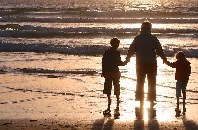 Three people, an adult and two children, stand hand in hand on a beach at sunset, their silhouettes reflected on the wet sand—capturing the essence of stress-free travel with children as waves roll in under a golden sky.