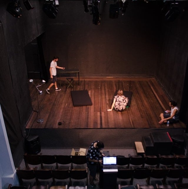 A small, empty theatre stage with three people rehearsing—a scene often seen in drama classes—one stands on the left, one sits floor centre, another sits right; a fourth person operates equipment at a table in the audience.