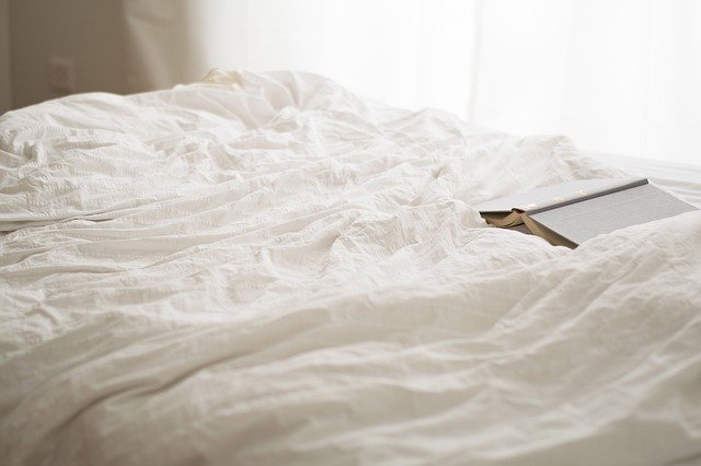 A neatly made bed with white rumpled sheets and a closed book resting on top, set near a window with sheer curtains letting in soft natural light—an inviting modern master bedroom perfect for exploring new modern colour ideas.