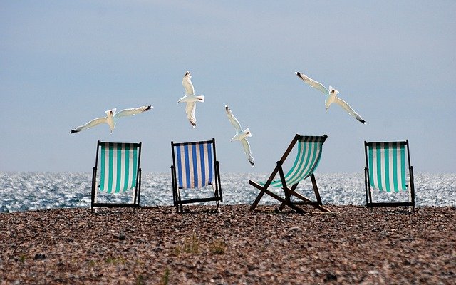 Four striped deckchairs sit on a pebbled beach, facing the sea, with five seagulls flying overhead—a perfect scene for family holidays and relaxing moments by the seaside.