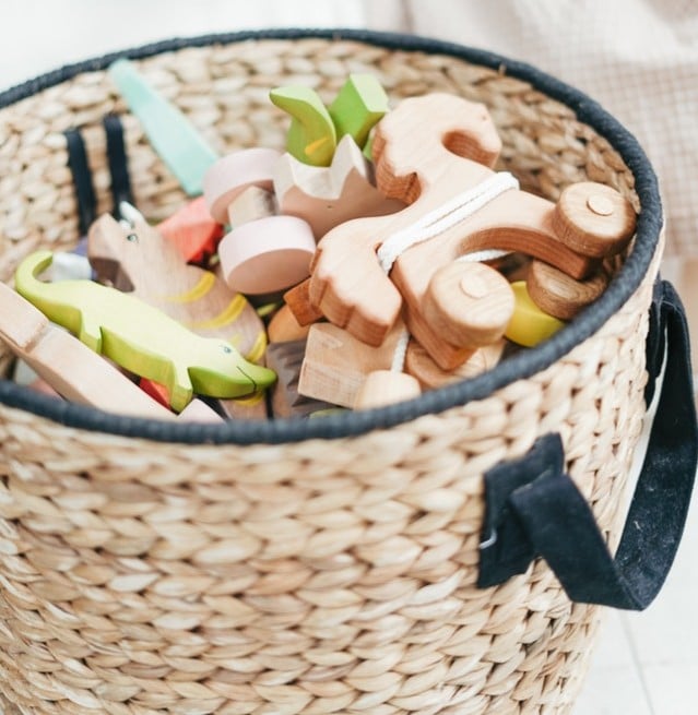 A woven basket with black handles, filled with wooden animal shapes and vehicles, sits on a light-coloured floor—ideal for playroom organisation and keeping your space tidy.