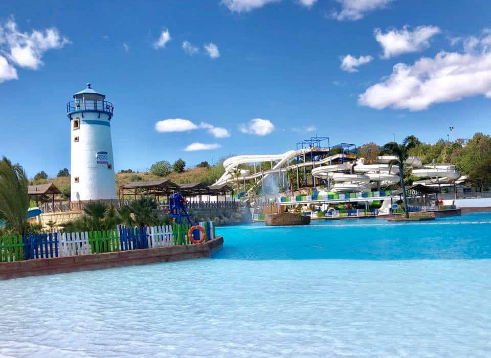 A bright water park scene in Benidorm features a blue and white lighthouse, water slides, a splash pad, and a large pool under a sunny sky. Green trees and a colourful fence surround this perfect family holiday destination.