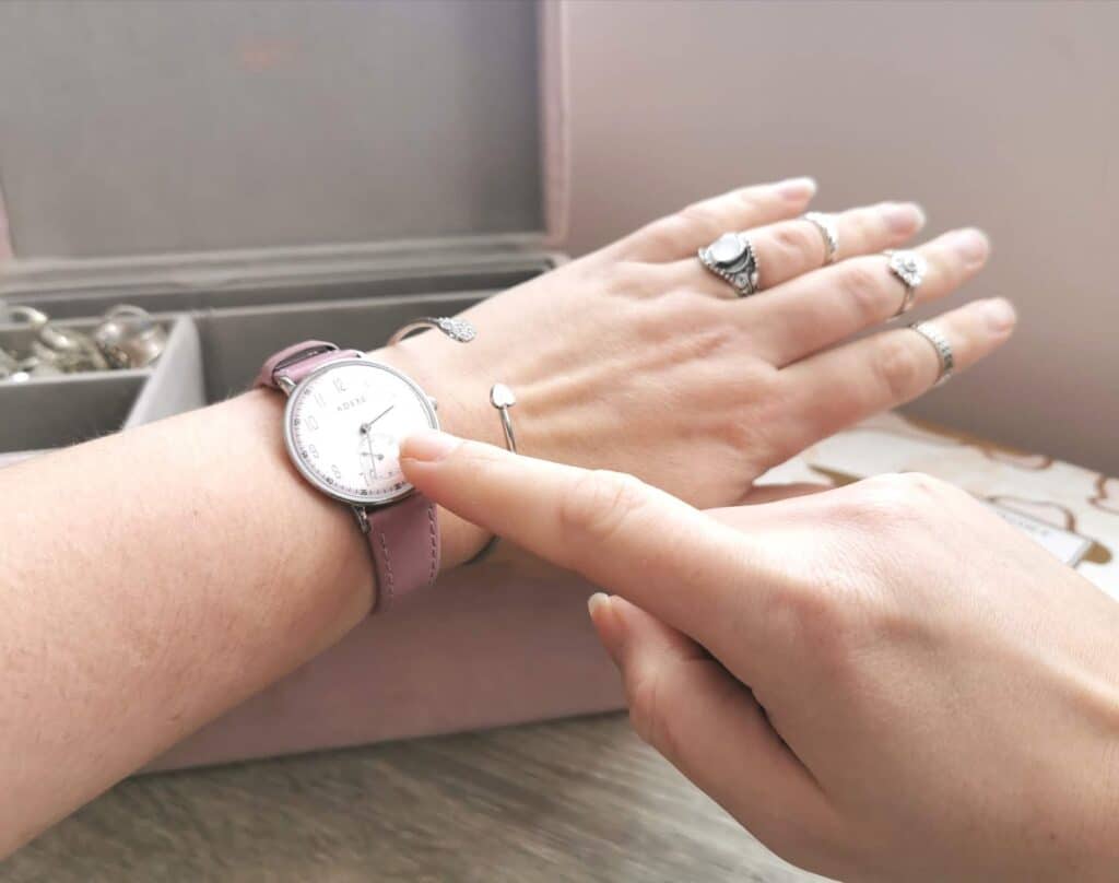 A hand with multiple rings points to the face of a wristwatch with a pink strap, worn on another hand—capturing the modern impatience in society. A jewellery box with rings is visible in the background.