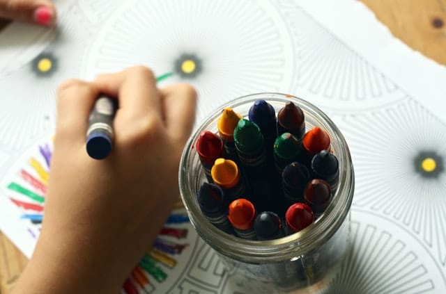A close-up of a hand colouring a patterned sheet with crayons, using a blue crayon. An open jar filled with colourful crayons is in the foreground—much like searching for the right childcare, each colour offers a unique choice for creativity.
