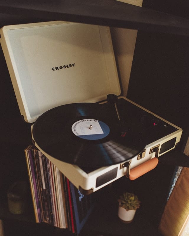 A white Crosley portable record player with a black vinyl spinning, placed on a shelf above a row of vinyl records and a small potted succulent—perfect for adding retro vibes to your man cave essentials.