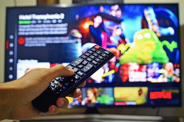 A hand holds a TV remote control pointed at a television screen displaying a streaming service interface, reflecting our impatient nation’s craving for instant access to countless colourful thumbnails and titles.