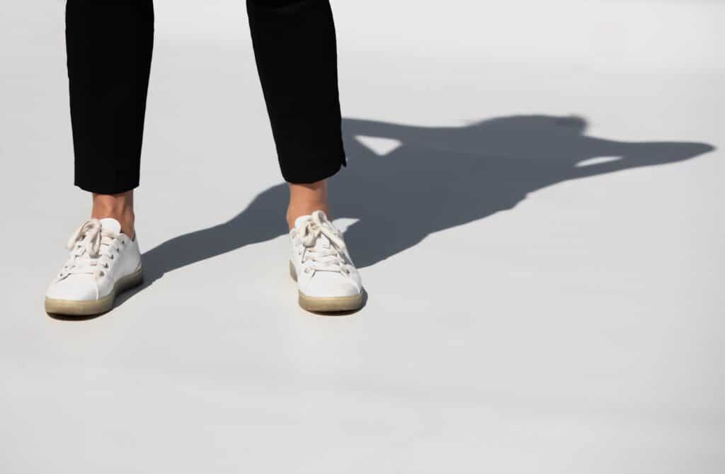 A person showcases women's trouser styles in black trousers and white trainers, standing on a white surface with a sharp shadow cast to the right. Only the lower legs and feet are visible.