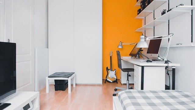 Modern tween to teen boys' bedroom featuring a bold orange feature wall, white furniture, desk with computer and lamp, wall shelves, guitar, TV, and a bed with chequered duvet on light wood flooring—perfect for a kids' room makeover.