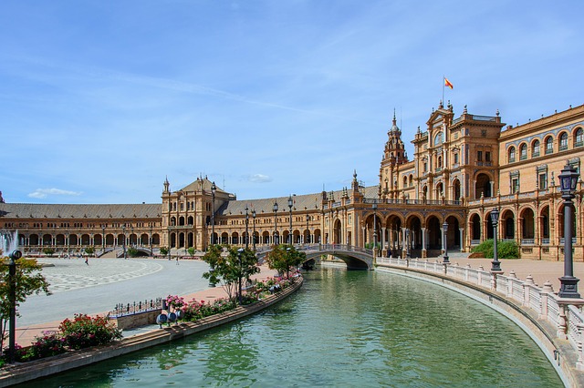 A grand, curved building with ornate towers and arches stands beside a canal with clear water and a small bridge; the Spanish flag flies atop under blue skies. Plaza de España in Seville is perfect for family activities and exploring Seville with children.