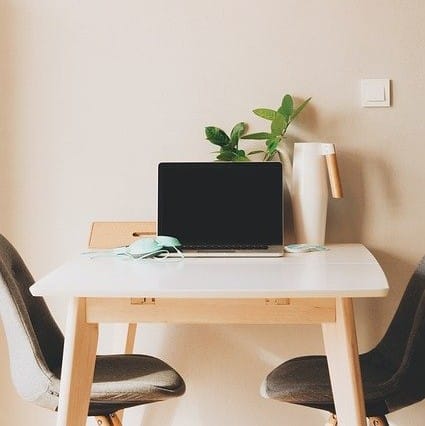 Minimalist workspace perfect for a boys' bedroom makeover, featuring a laptop on a white desk between two grey chairs. A green plant, white vase, and face mask add style to this tween to teen space against a light beige wall.
