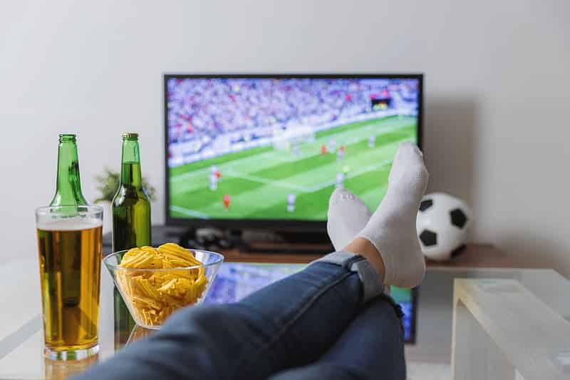 A person with feet up on a table in a cosy man cave watches a football match on TV, surrounded by two beer bottles, a glass of beer, crisps, and a football—the perfect setting for enjoying classic man cave benefits.