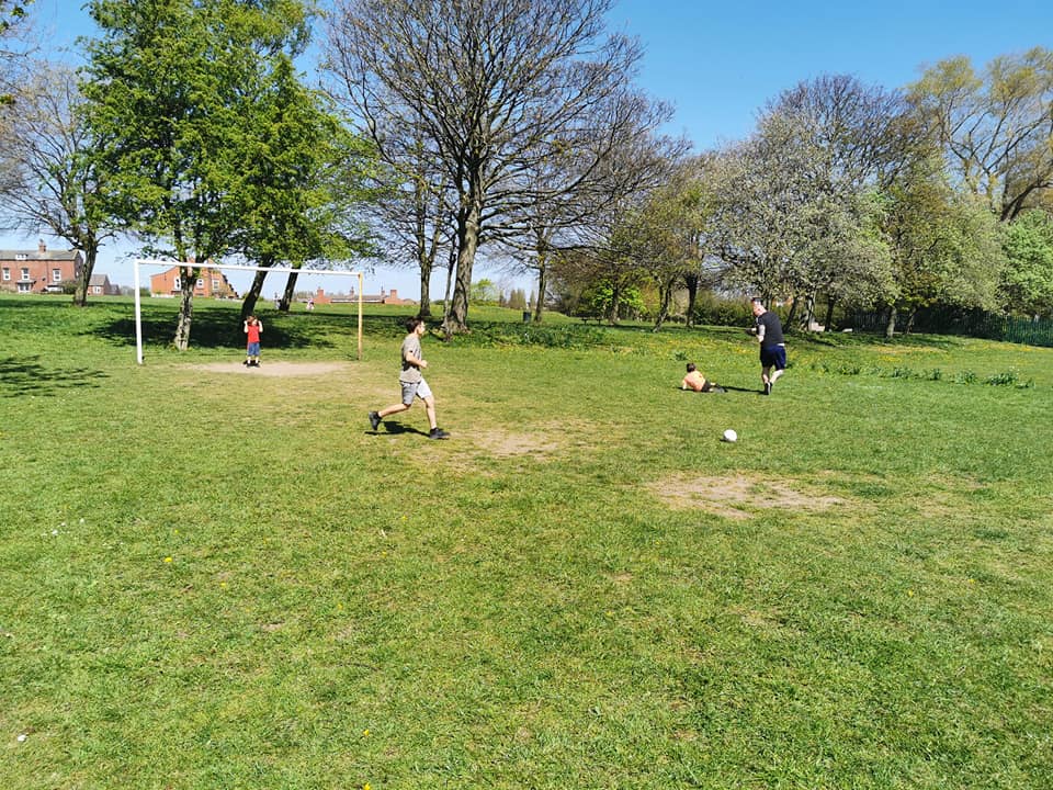 Four children enjoy football on a grassy field surrounded by trees and houses, showing fun ways to exercise outdoors on a sunny day. One runs towards the ball while others scatter, demonstrating simple daily exercise for kids.